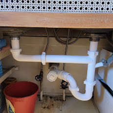 White PVC plumbing pipes under a sink with valves and a red bucket nearby.